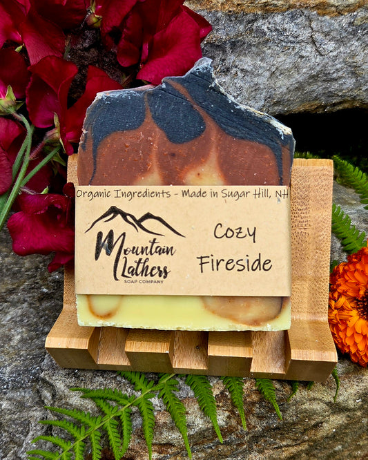 A handmade soap bar displayed on a small wooden stand outdoors, with a paper label reading Mountain Lathers Cozy Fireside. The soap has a marbled top with dark charcoal, rust orange, and brown tones, and a pale yellow base.