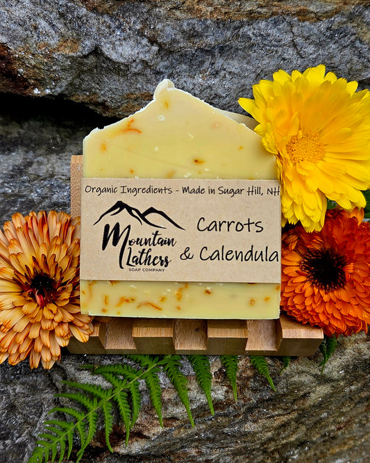 A handmade bar of soap labeled 'Carrots & Calendula' by Mountain Lathers, displayed on a small wooden stand with orange and yellow flowers and green ferns against a rustic rock background.