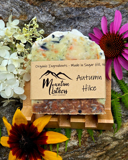 A handmade soap bar labeled 'Autumn Hike' by Mountain Lathers, styled with flowers and greenery on a wooden stand against a stone background.