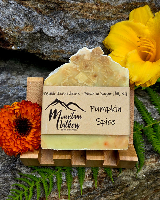 A handmade bar of soap labeled 'Mountain Lathers Organic Ingredients - Made in Sugar Hill, NH' and 'Pumpkin Spice,' displayed on a small wooden stand. The soap has a rustic irregular top and pale warm yellow-orange color.