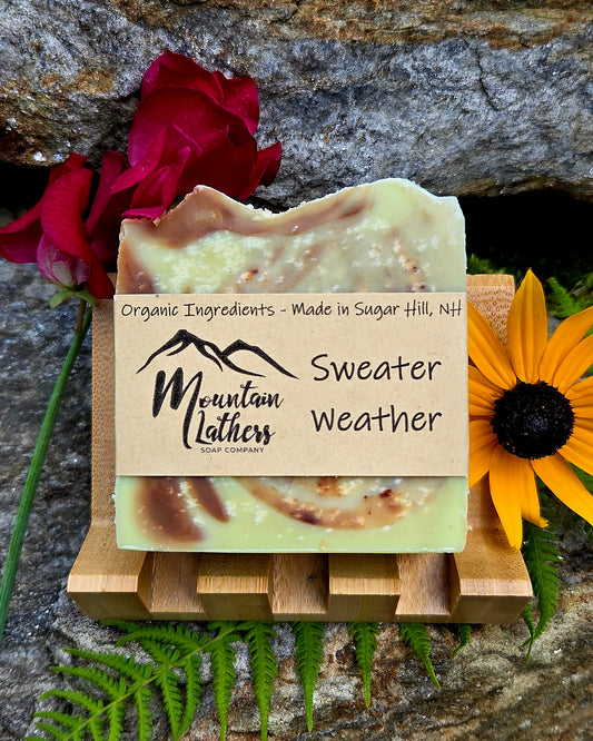 A handmade soap bar labeled 'Sweater Weather' by Mountain Lathers Hand Company, displayed on a small wooden stand. The soap has a pale yellow-green base with brown swirls and is styled with red roses, a yellow flower, and green ferns against a textured gray rock background.