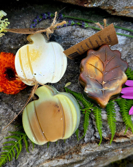 Three artisanal soap bars shaped like pumpkins and a leaf, styled with green ferns, orange flowers, and a small wooden tag on a textured gray stone background.