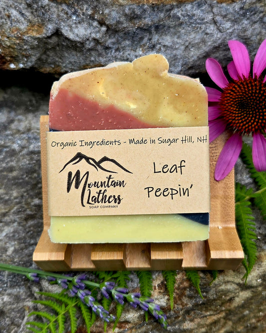 A handmade soap bar labeled 'Leaf Peepin'' by Mountain Lathers Soap Company, displayed on a small wooden stand. The soap has a marbled color story of yellow, pink, and pale green, with a kraft paper label wrapped around it.