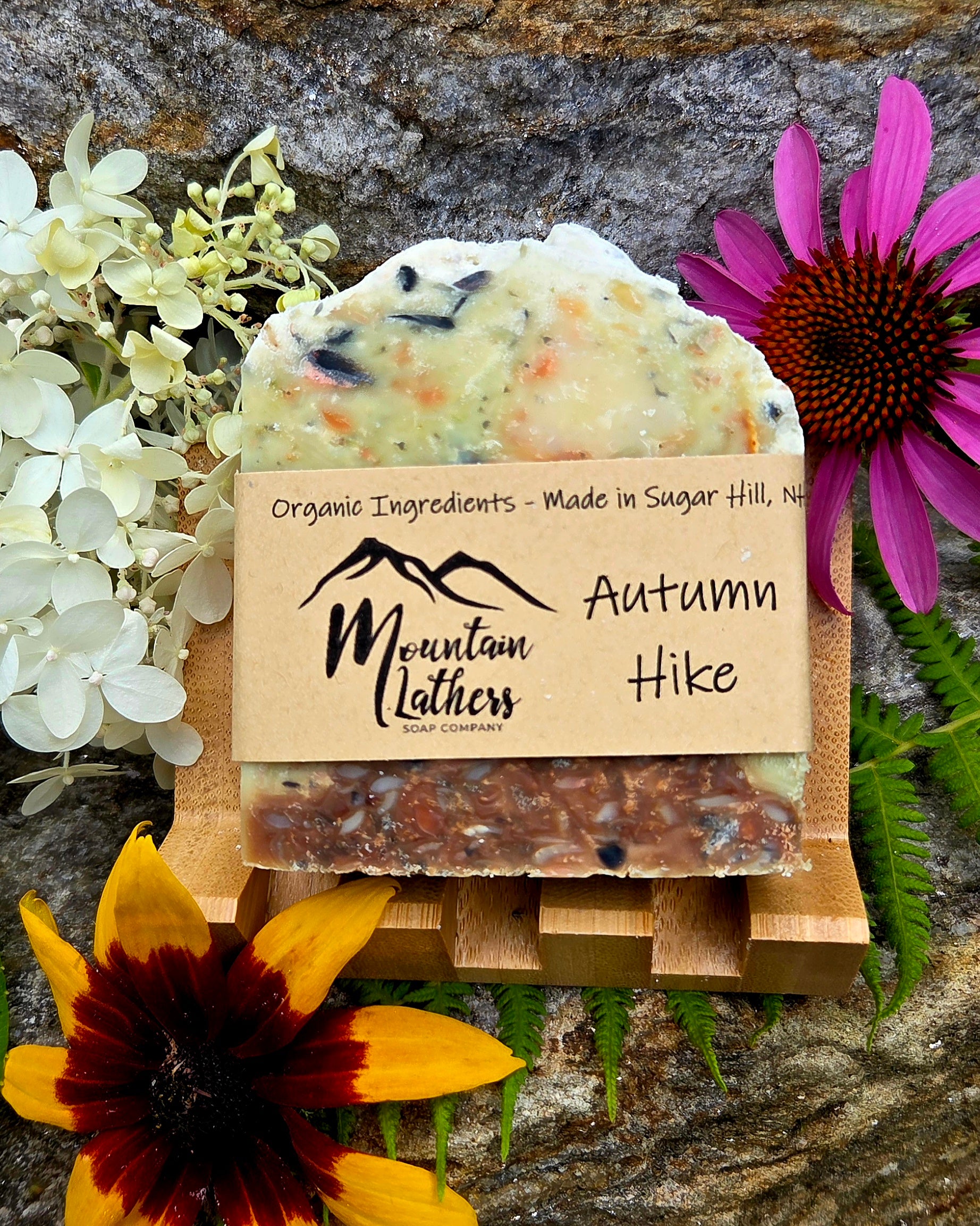 A handmade soap bar labeled 'Autumn Hike' by Mountain Lathers, styled with flowers and greenery on a wooden stand against a stone background.