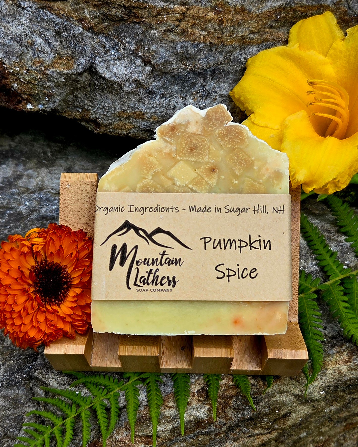 A handmade bar of soap labeled 'Mountain Lathers Organic Ingredients - Made in Sugar Hill, NH' and 'Pumpkin Spice,' displayed on a small wooden stand. The soap has a rustic irregular top and pale warm yellow-orange color.