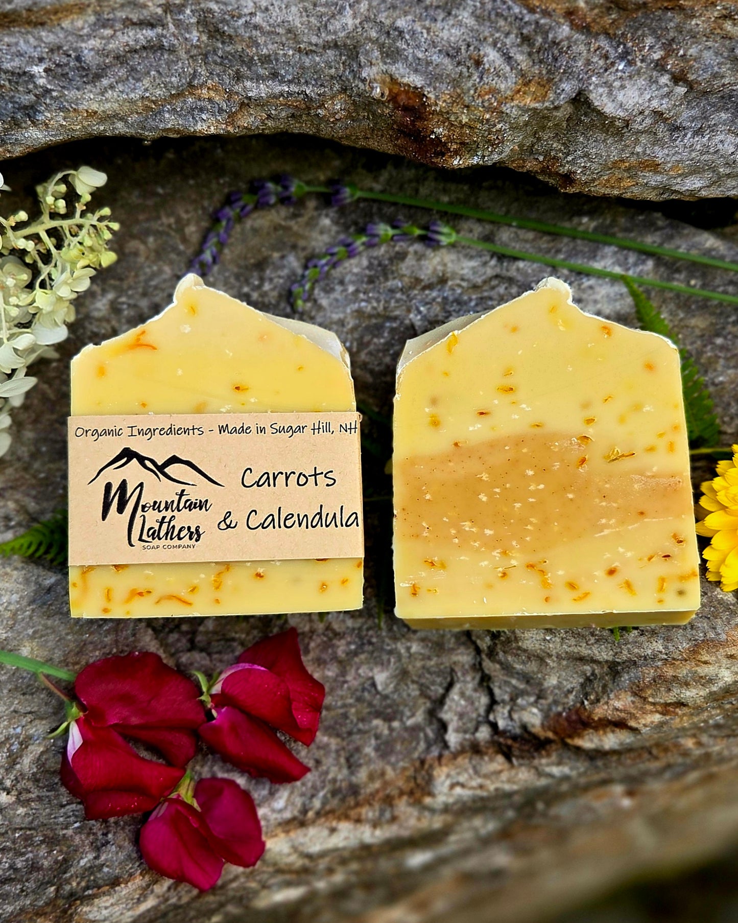 Two rectangular bars of handmade soap labeled 'Mountain Lather Carrots & Calendula' resting on a textured gray stone surface, surrounded by red rose petals, green stems, and small yellow and white flowers.