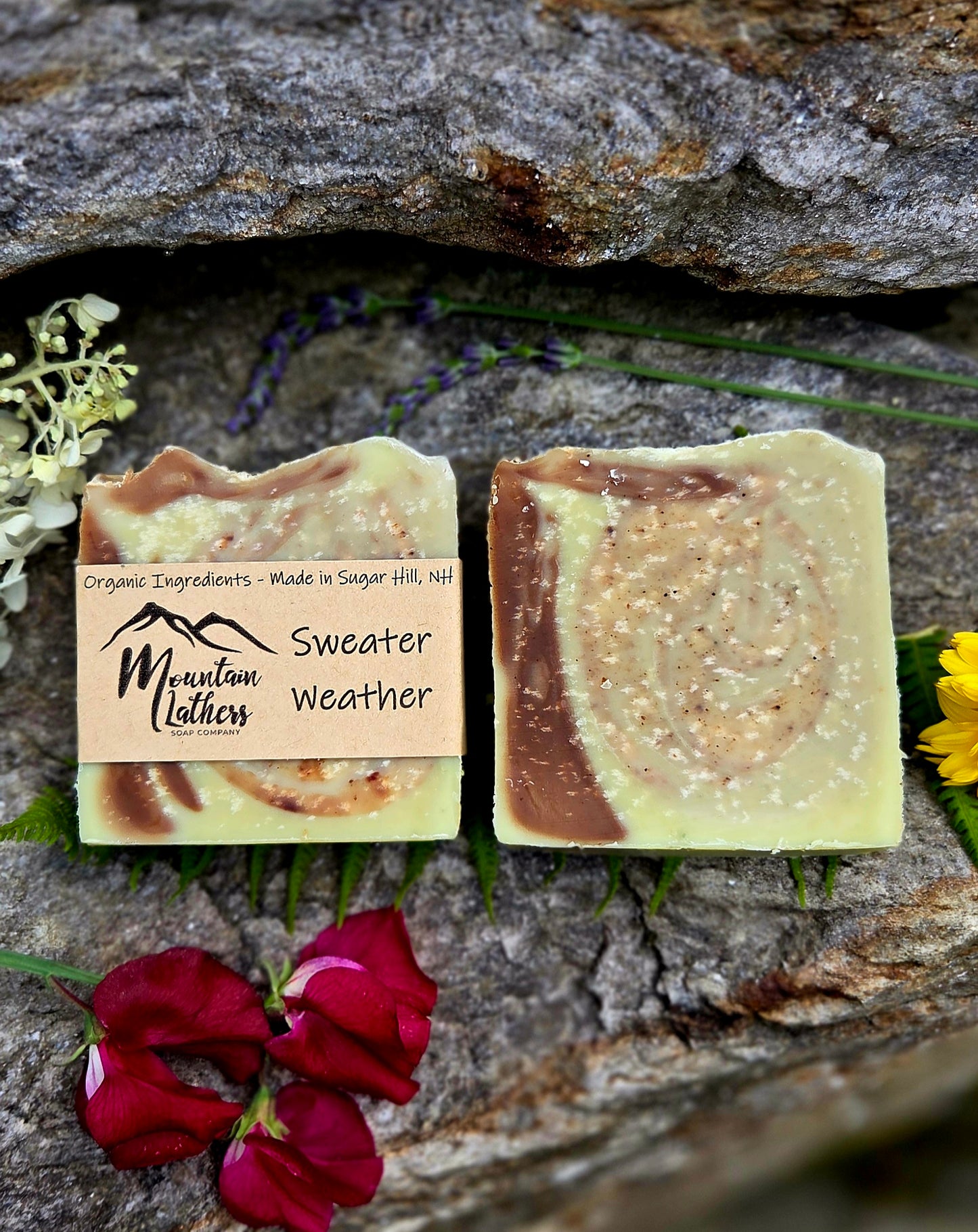 Two handmade soap bars with a swirled cream, brown, and pale yellow pattern, displayed on a textured gray rock with red flowers and green sprigs. One soap bar has a paper label that reads 'Mountain Lathers' and 'Sweater Weather'.
