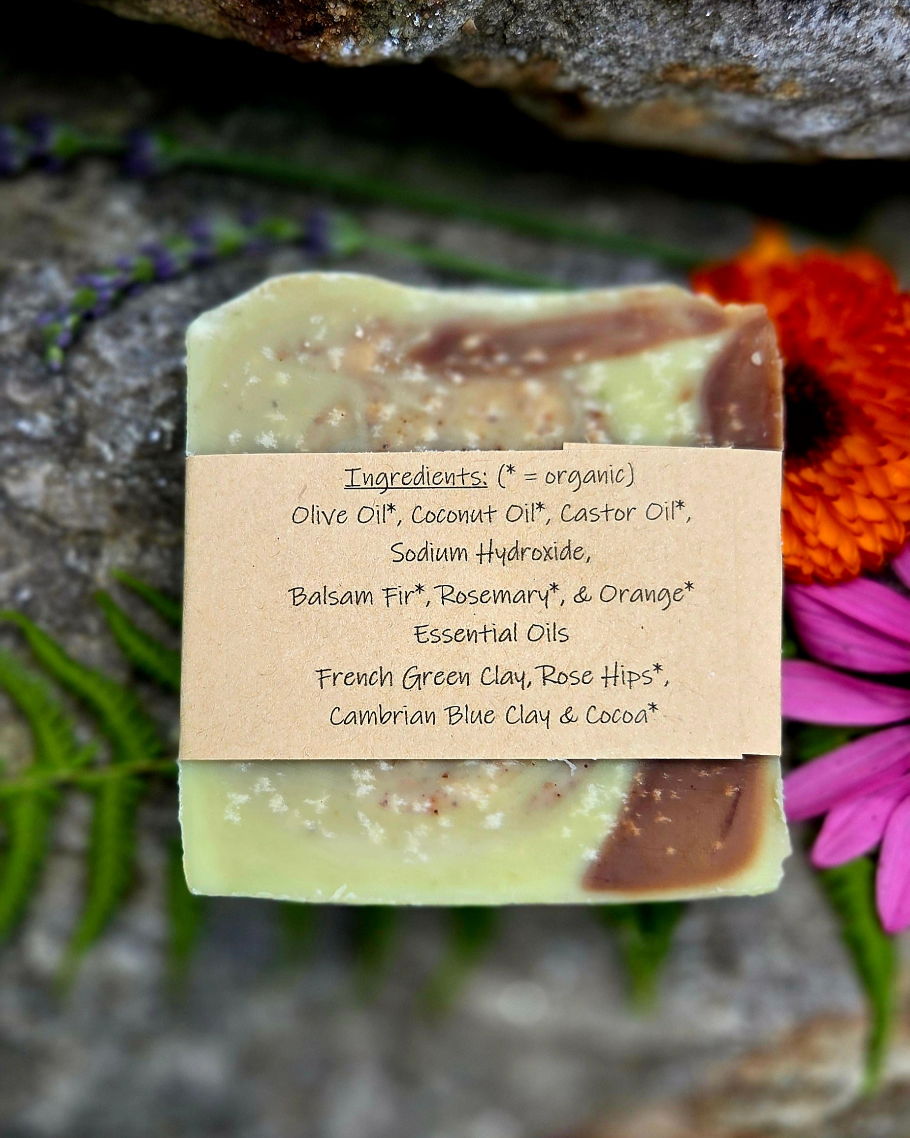 A handmade rectangular bar of soap with a kraft paper label showing ingredients, styled on a dark stone background with purple and orange flowers.