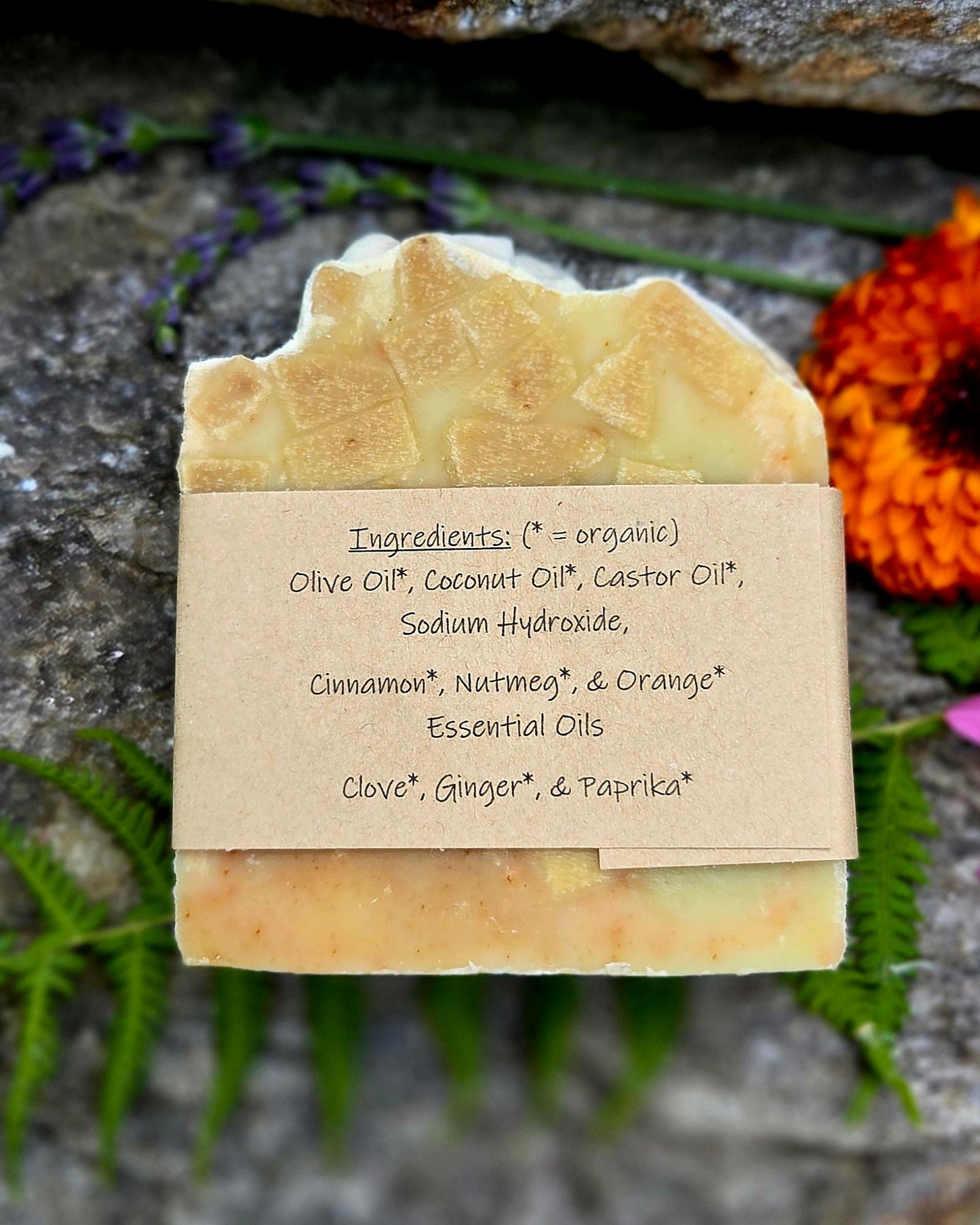 A handmade rectangular bar of soap with a rustic, uneven top edge, featuring a natural beige paper label with handwritten ingredients. The soap is styled on a dark textured stone surface with green ferns and purple flowers in the background.