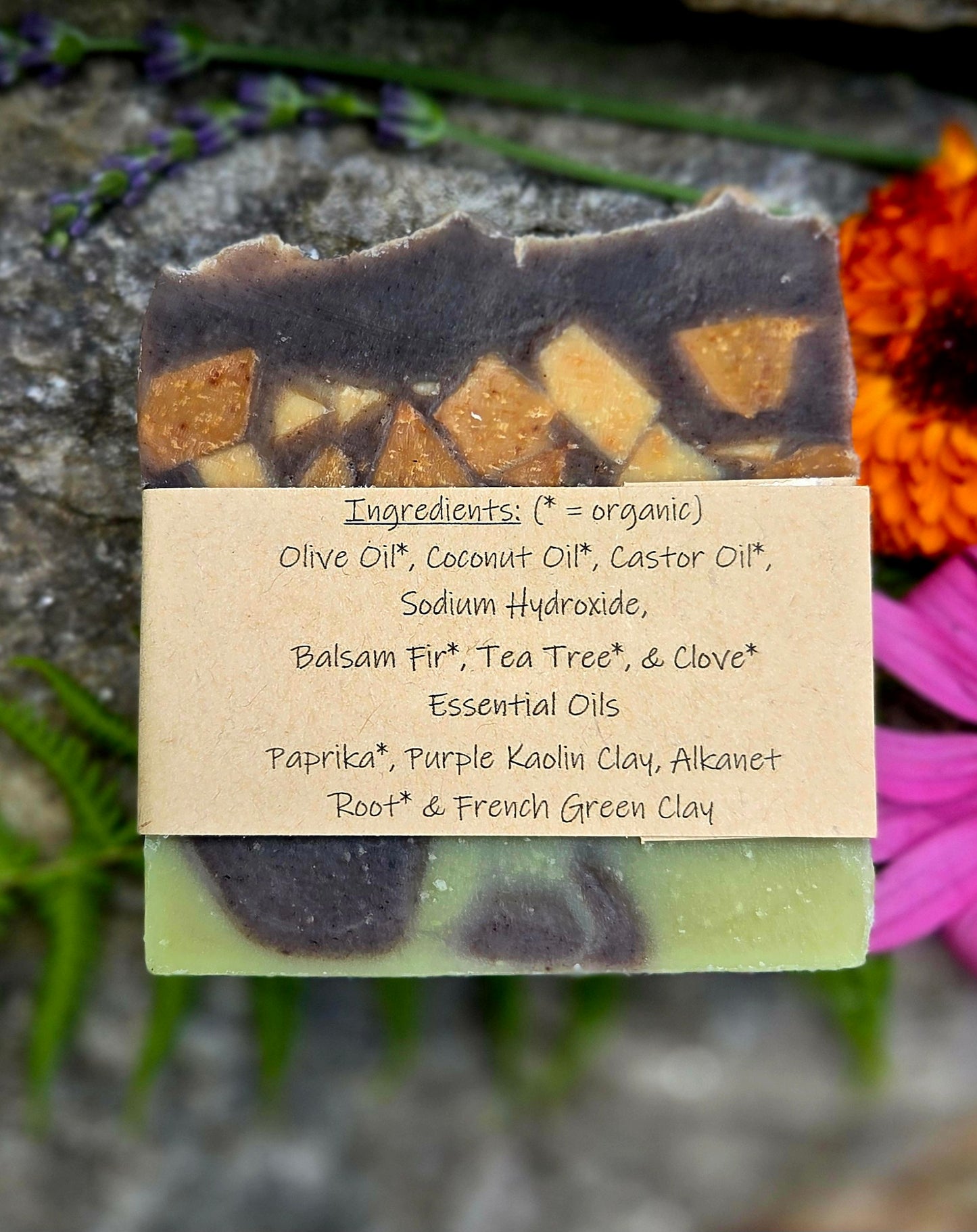 A handmade soap bar with a rustic irregular top edge, featuring a dark purple upper layer with embedded amber pieces and a pale green lower layer. A kraft paper label wrapped around the soap lists ingredients including olive oil, coconut oil, castor oil, sodium hydroxide, balsam fir, tea tree, clove, paprika, purple kaolin clay, alkanet, and root & French green clay.