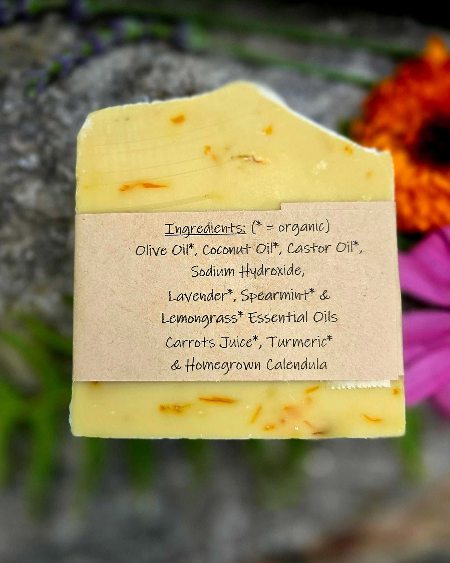 A rectangular bar of pale yellow soap with small orange botanical flecks, featuring a brown kraft paper label with handwritten ingredients. The background is a soft-focus outdoor scene with green foliage and orange flowers.