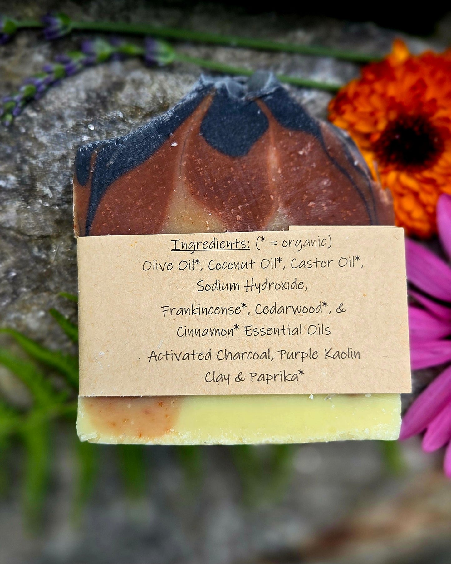 A handmade soap bar with a kraft paper label showing ingredients, set against a blurred natural background with flowers.