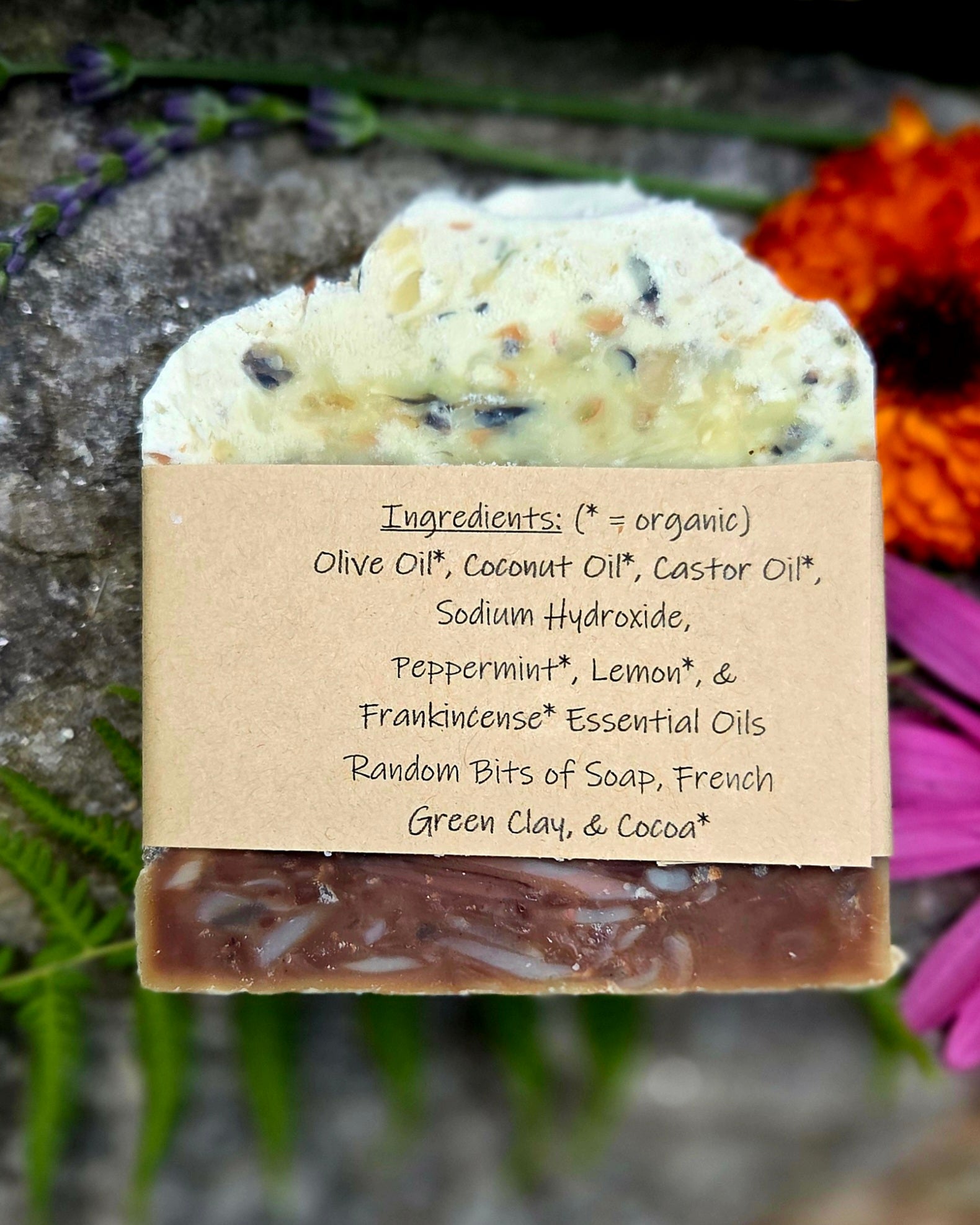 A handmade soap bar wrapped in a kraft paper label, displayed on a rustic stone surface with flowers and greenery in the background. The label lists ingredients including olive oil, coconut oil, castor oil, sodium hydroxide, peppermint, lemon, and green clay.