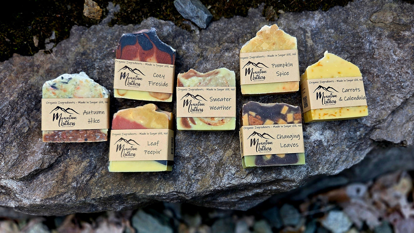 Six handmade bars of soap from Mountain Lathers Soaps are arranged on a dark, textured rock outdoors. Each bar has a unique earthy color palette and a paper label with the brand name 'Mountain Lathers Soaps' and a mountain logo.