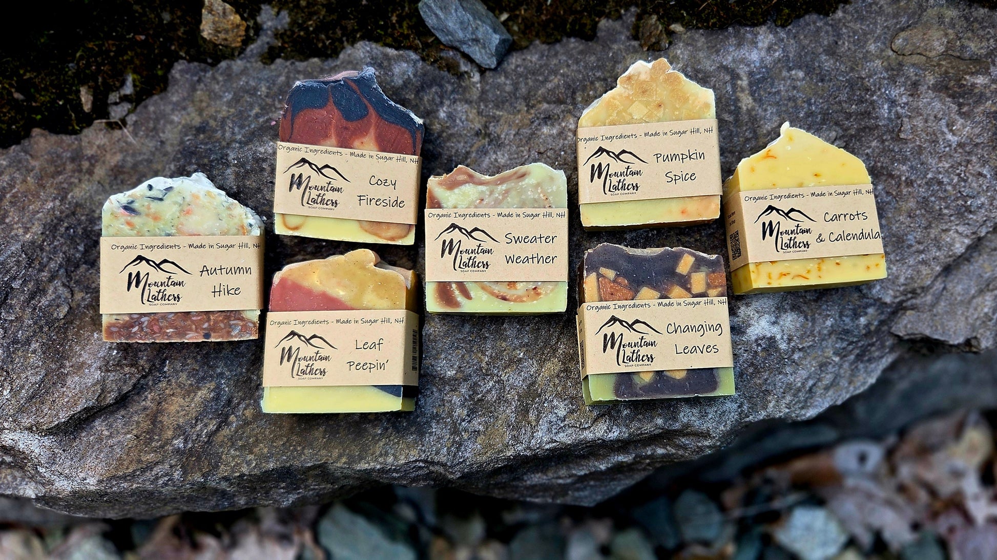 Six handmade bars of soap from Mountain Lathers Soaps are arranged on a dark, textured rock outdoors. Each bar has a unique earthy color palette and a paper label with the brand name 'Mountain Lathers Soaps' and a mountain logo.