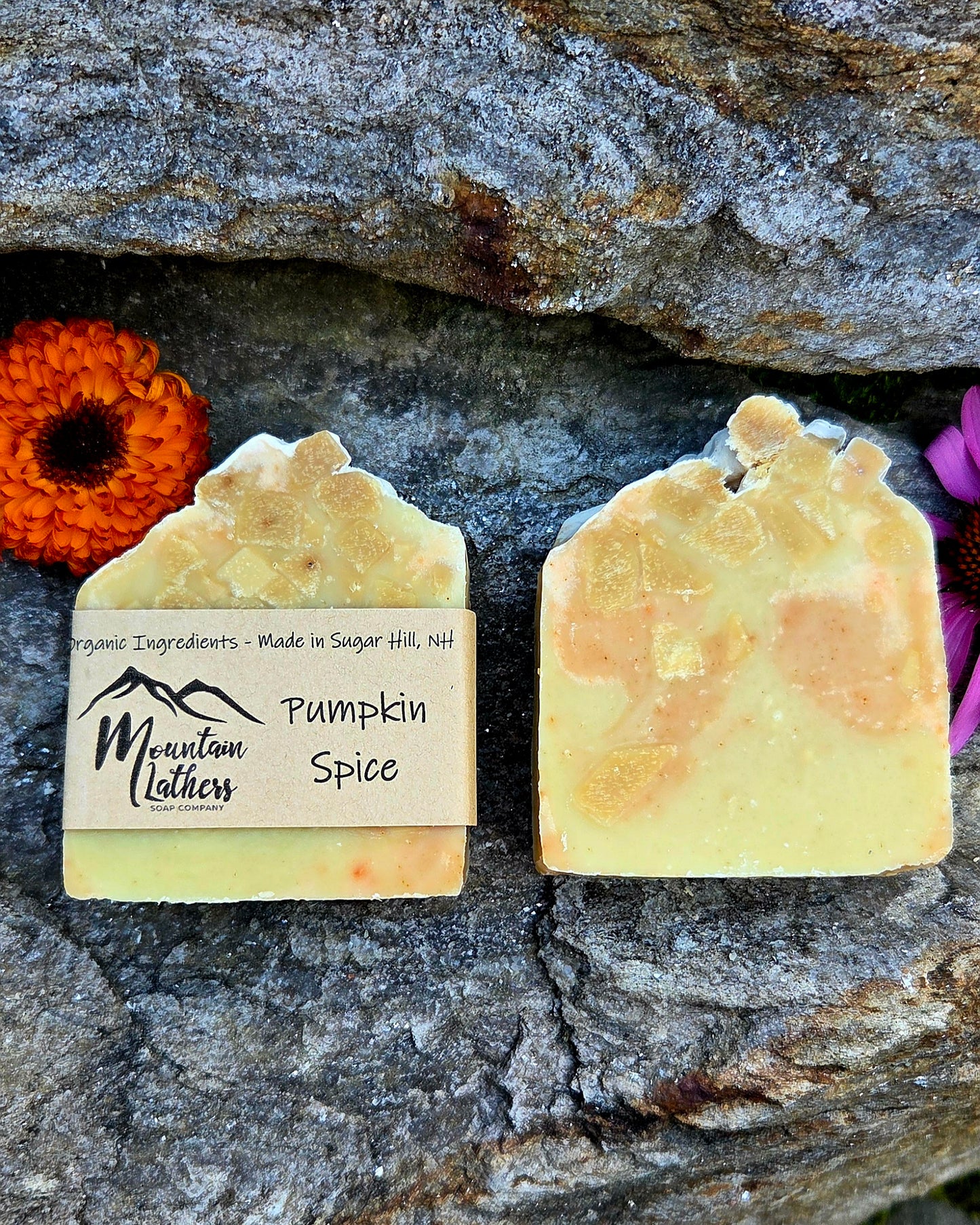 Two handmade soap bars labeled 'Pumpkin Spice' by Mountain Lathers, displayed on a textured gray stone background with decorative orange and purple flowers.