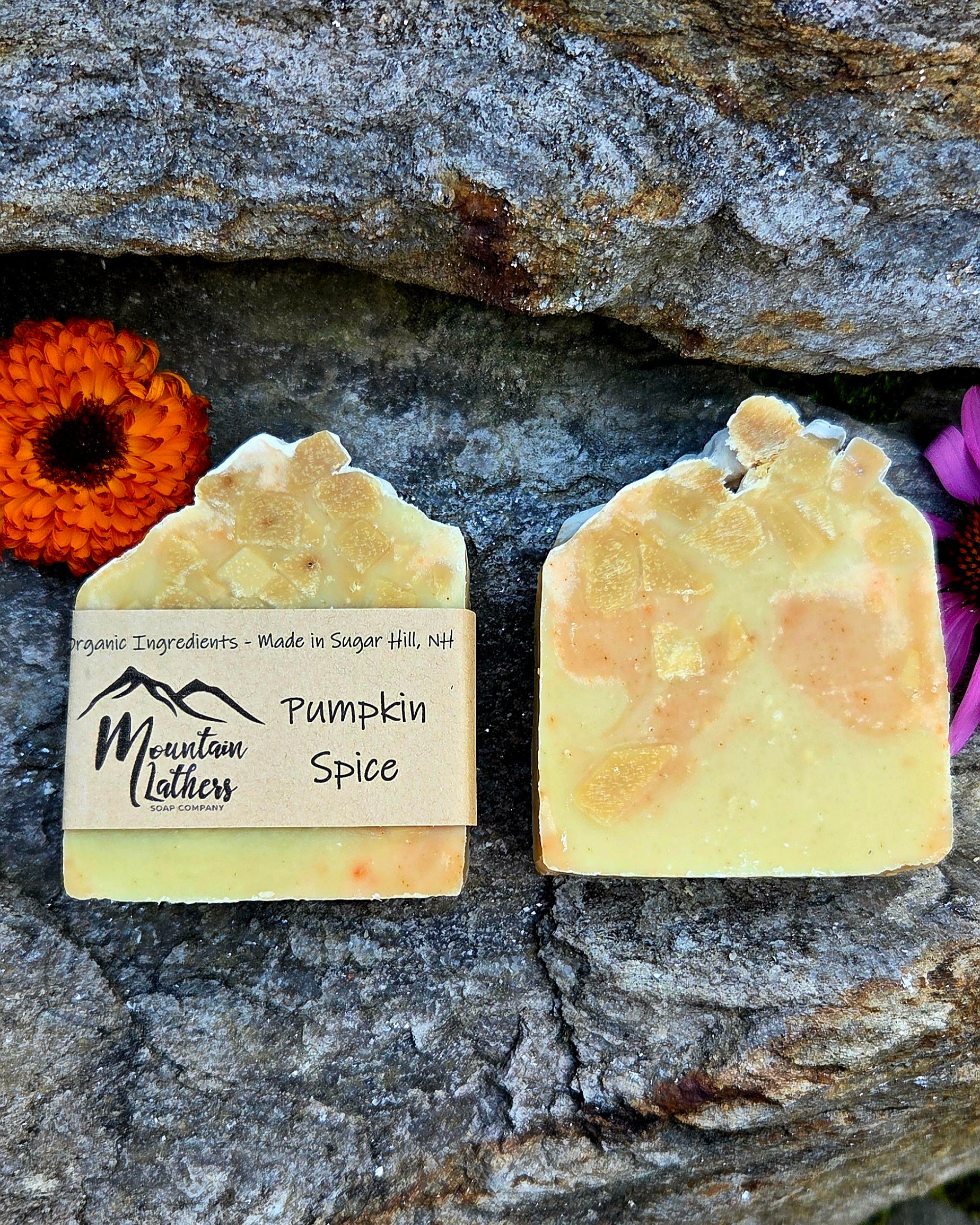 Two handmade soap bars labeled 'Pumpkin Spice' by Mountain Lathers, displayed on a textured gray stone background with decorative orange and purple flowers.